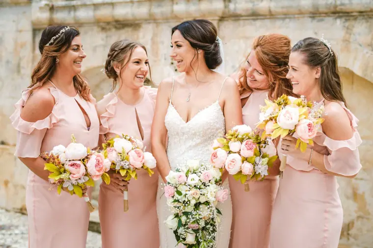Bride laughing with bridesmaids in blush dresses holding pastel bouquets