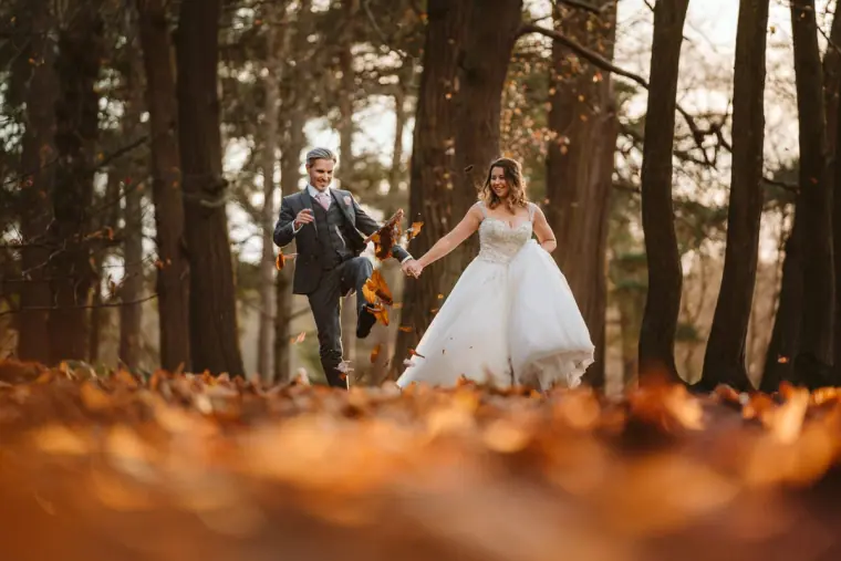 Bride laughing as the groom kicks up autumn leaves in a pine wood