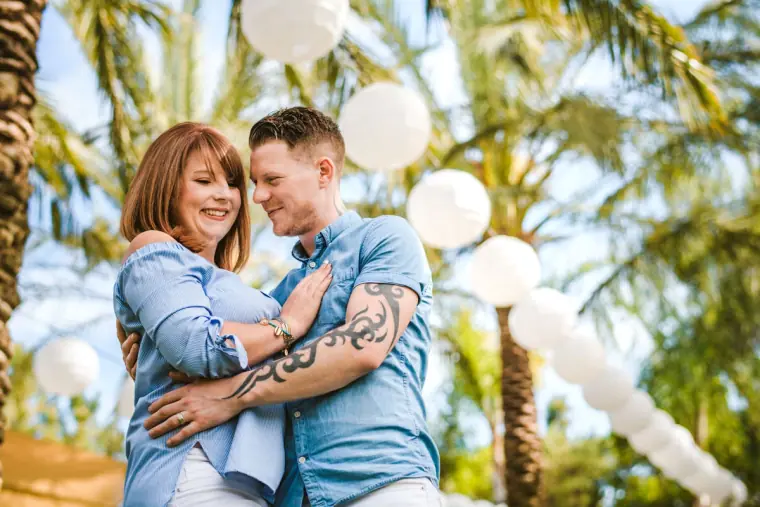 Couple in matching blue shirts embracing beneath palms and white balloons