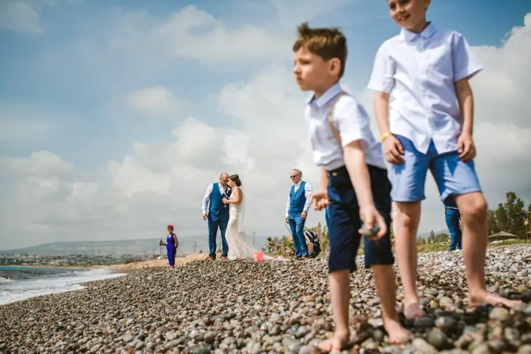 Two page boys on a pebble beach as the bride and groom stand by the sea