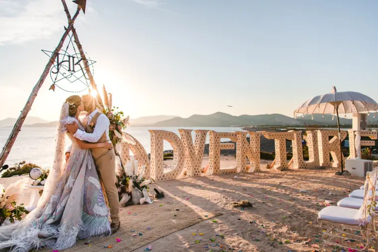 Couple kissing beside giant ADVENTURE letters and a monogrammed tipi frame by the sea