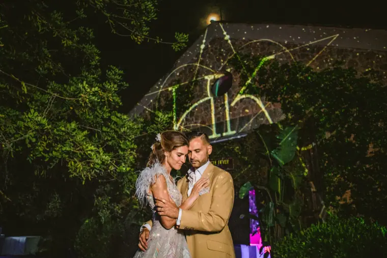 Couple embracing beneath a glowing neon monogram among trees at night
