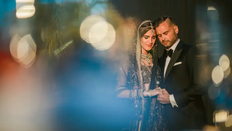 Bride in an embellished veil and groom in a tuxedo, framed by blue bokeh