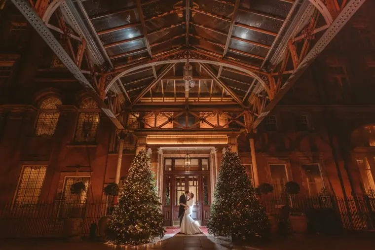 Couple at the end of a grand glass-roofed hall lined with Christmas trees