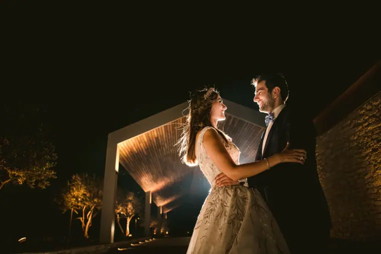 Bride and groom facing each other at night beside a lit modern pergola