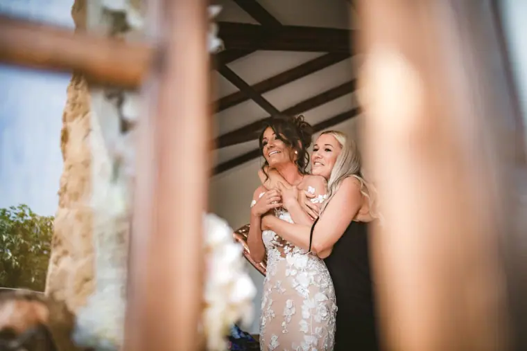 Bride hugging a bridesmaid, framed by wooden beams and stone