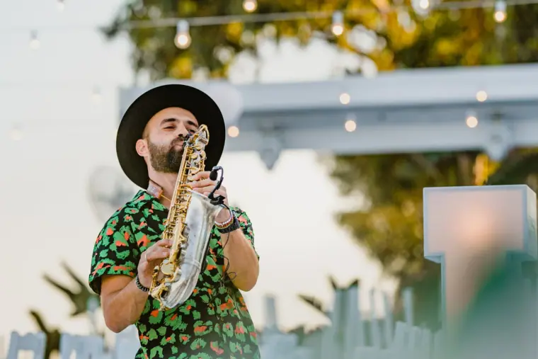 Saxophonist in a hat and patterned shirt playing outdoors under festoon lights