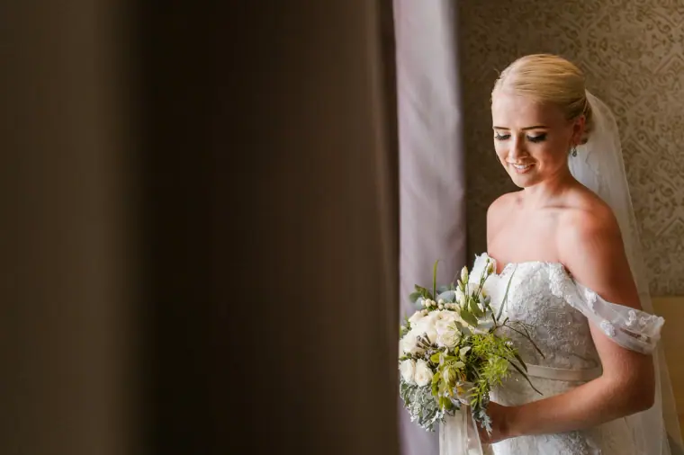 Bride in an off-the-shoulder gown holding a white bouquet by a window