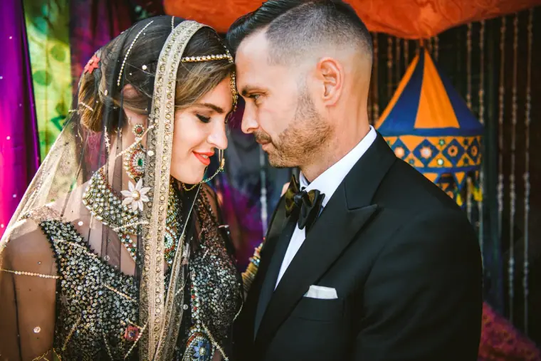 Bride in a jewelled dupatta and groom in a tuxedo touching foreheads