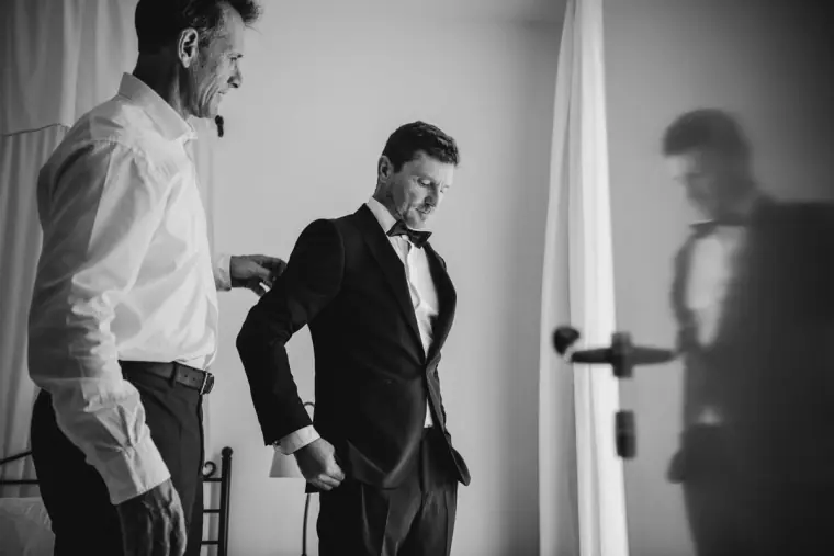 Black-and-white photo of the groom in a tuxedo being helped into his jacket