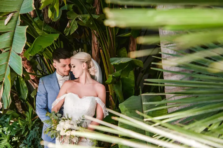 Couple embracing among tropical leaves, she holds a pale bouquet