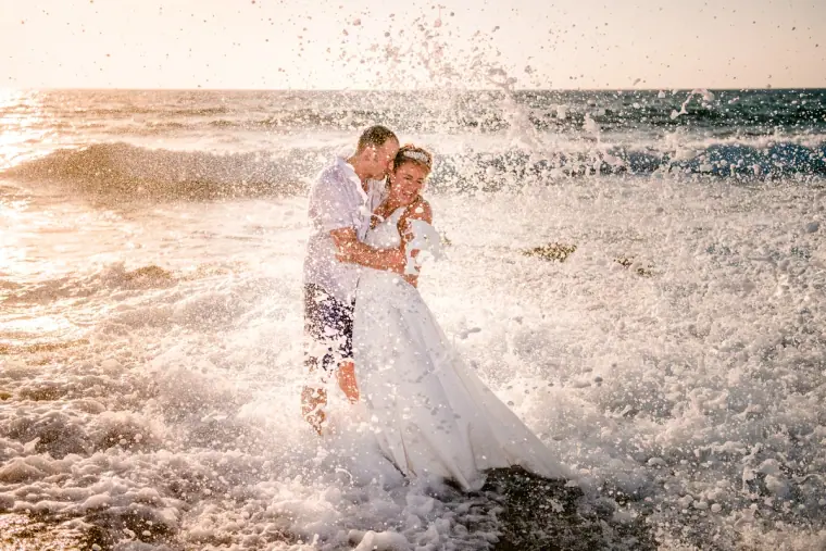 Groom carrying the bride as a wave crashes behind them