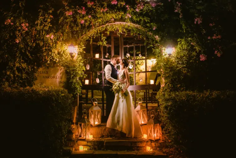 Couple kissing beneath a flower-covered arch lit by lanterns at night