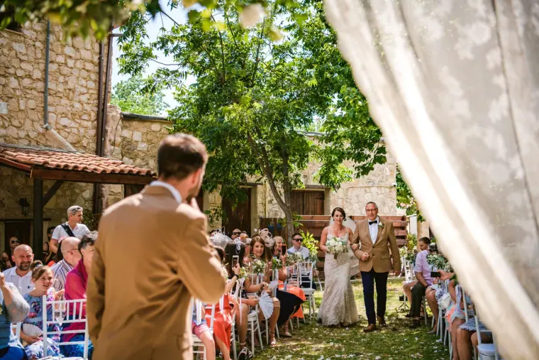 Bride walking down a garden aisle with her father as the groom waits by a curtain