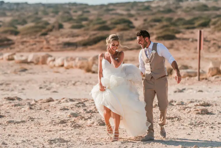 Bride lifting her dress and laughing as the groom walks beside her on sand