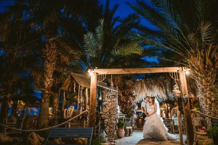 Couple beneath a rustic lantern-lit pergola among palms at dusk