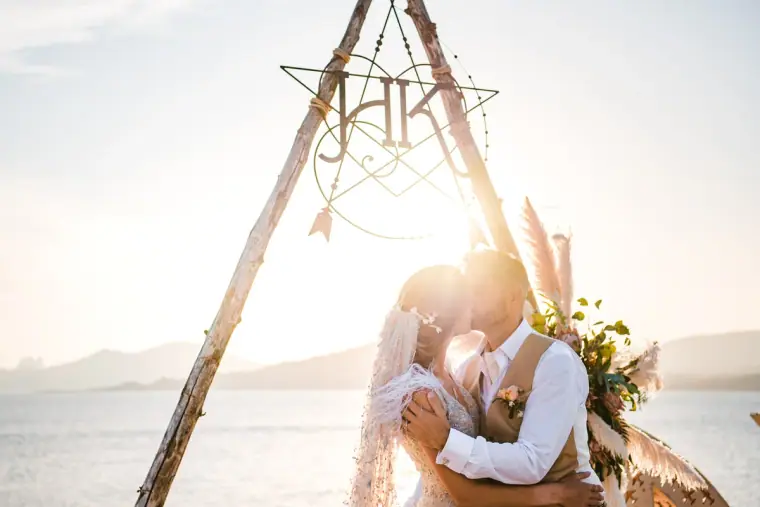 Couple kissing beneath a wooden triangle arch with a dreamcatcher, sun flaring over the sea