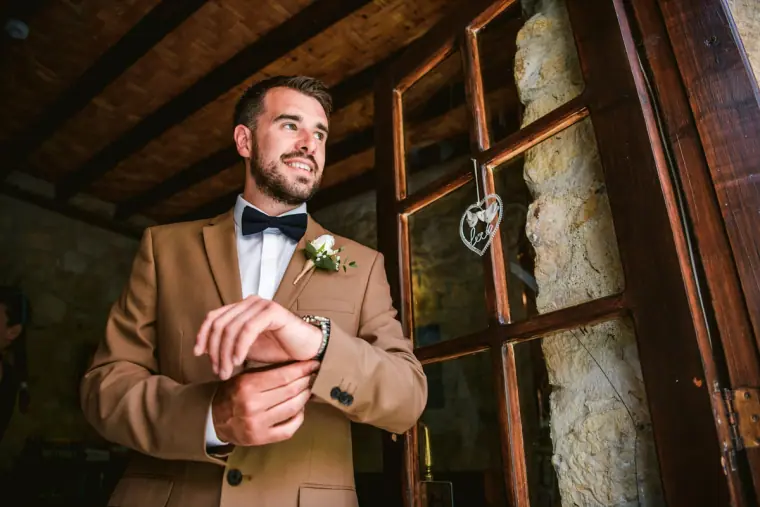 Smiling groom in a tan suit and bow tie adjusting his cuff by a wooden doorway