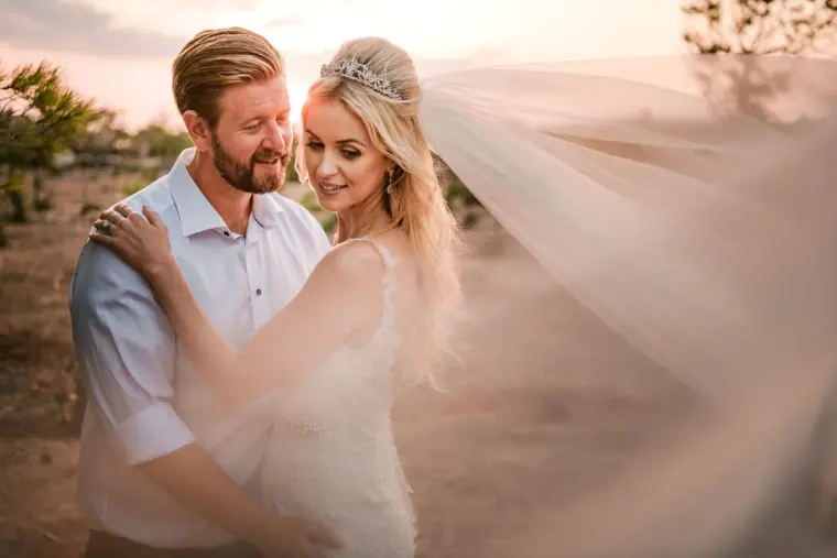 Groom holding his smiling bride as her long veil billows in golden evening light