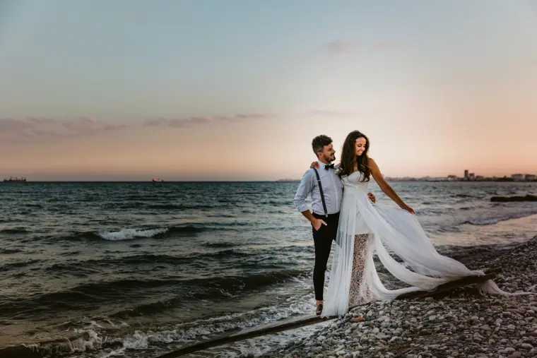 Bride and groom walking along a pebbly shoreline at dusk, her sheer skirt blowing in the breeze
