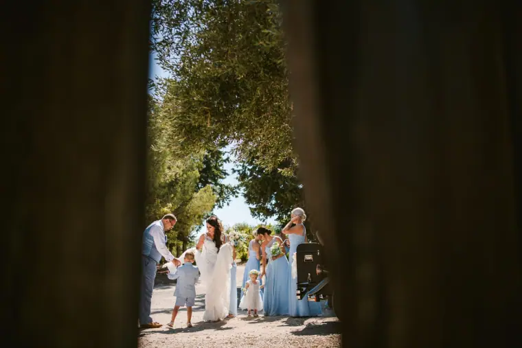 Bride and her bridesmaids in pale blue seen through a gap in dark curtains, among olive trees