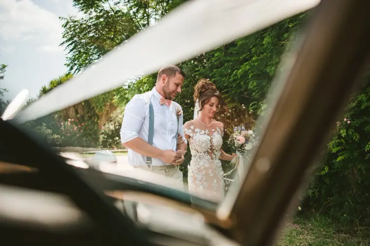 Bride and groom framed through a vintage car window as they stand among greenery