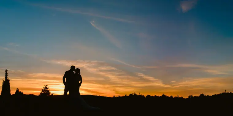 Silhouette of the couple embracing on a hilltop against a blue and orange sunset sky