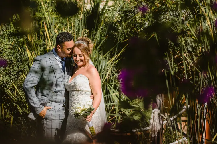 Groom in a checked suit kissing his bride's cheek among lush garden plants