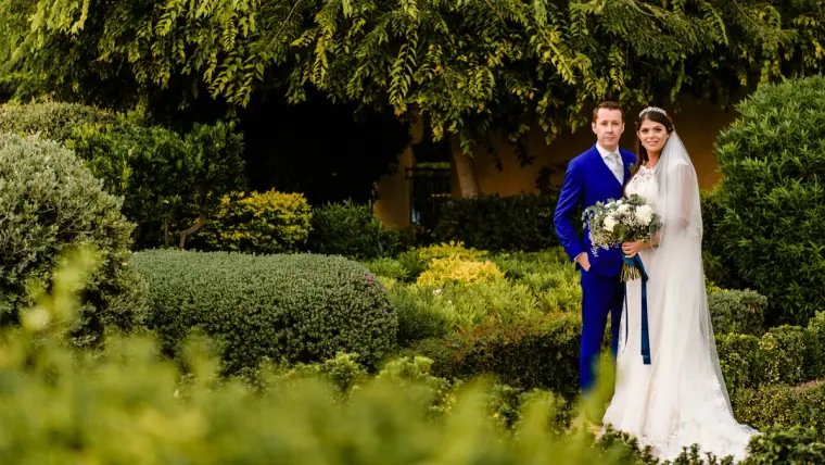 Groom in a royal blue suit and bride with her bouquet standing in a manicured garden