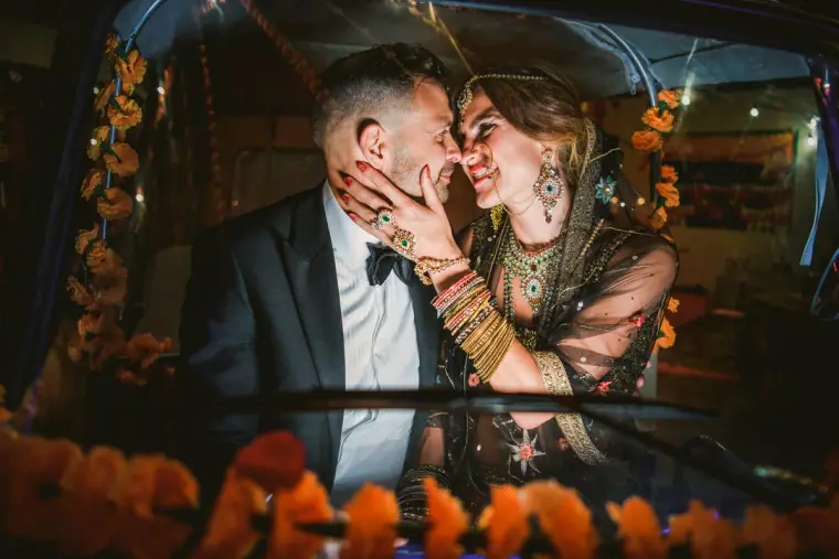 Bride in Indian jewellery cupping the groom's face inside a car decorated with marigolds