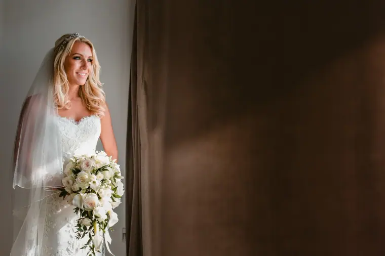 Bride in a strapless gown and veil holding white roses beside a brown curtain