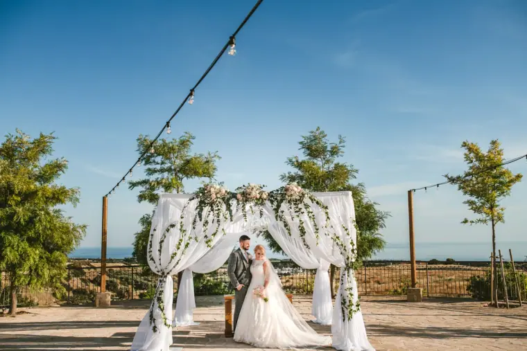 Bride and groom beneath a white floral ceremony canopy with festoon lights and sea view