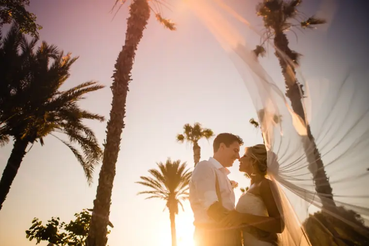 Couple embracing between tall palm trees at sunset, her veil catching the light