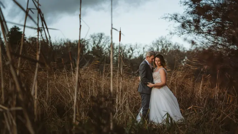 Bride and groom standing among tall dry reeds and grasses under a cloudy sky