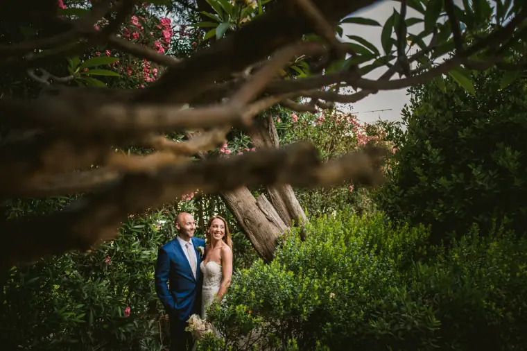 Bride and groom framed by branches and pink flowers in a leafy garden