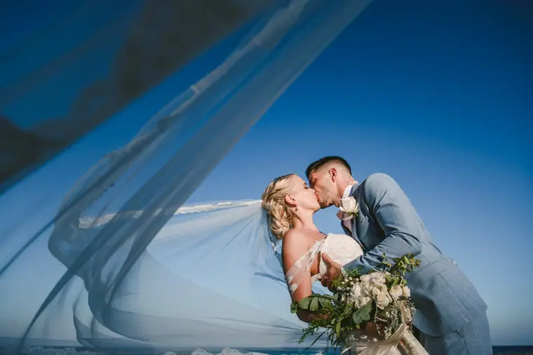 Bride and groom kissing against a blue sky with her veil sweeping across the frame