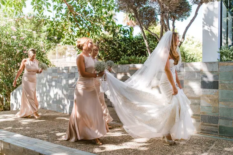 Bridesmaids in champagne dresses helping the bride with her long veil on a sunny terrace