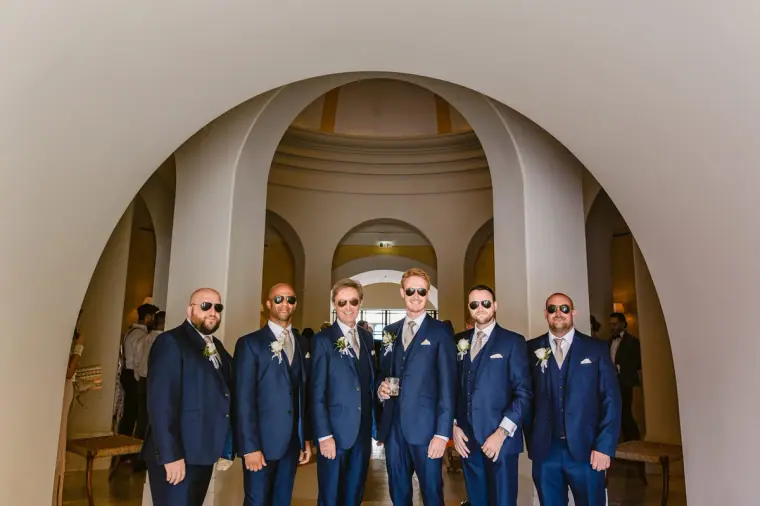 Groom and groomsmen in navy suits and sunglasses lined up under a white arch