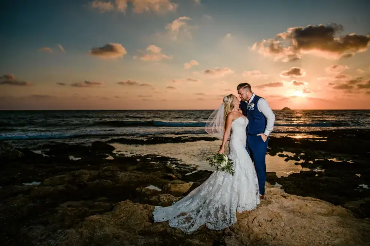 Bride and groom standing on dark rocks by the sea at sunset