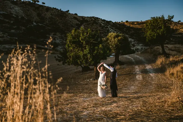 Groom twirling his bride on a dry hillside path among olive trees
