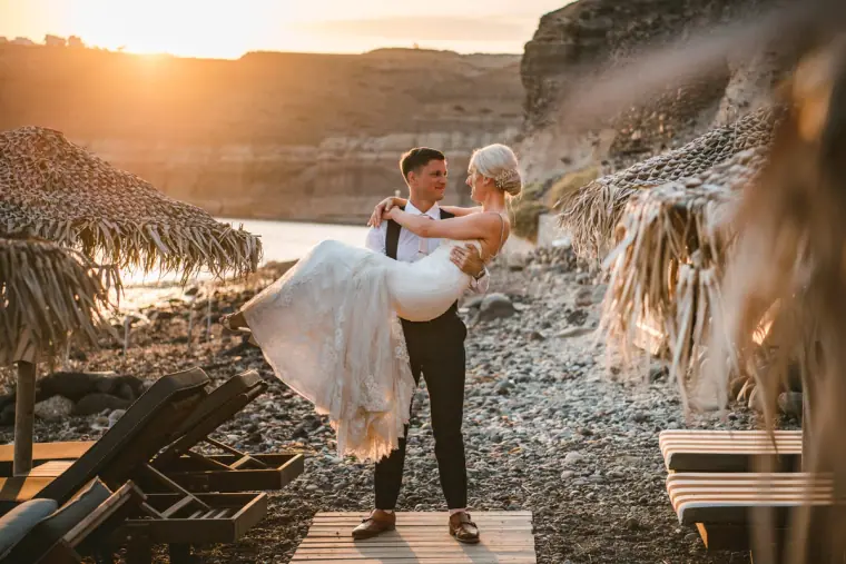 Groom carrying his laughing bride on a pebble beach beside thatched parasols at sunset