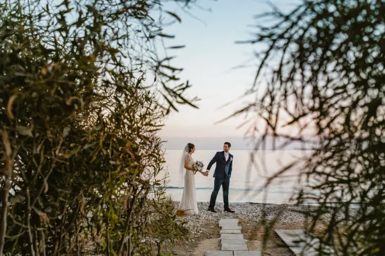 Couple holding hands by the sea, framed by feathery bushes