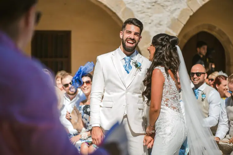 Groom in a cream suit laughing beside his bride as guests applaud at the ceremony