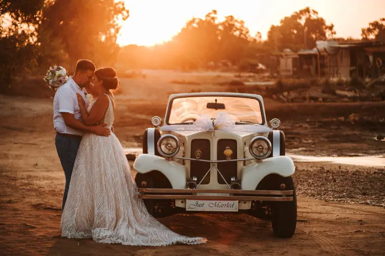 Bride and groom kissing beside a vintage white wedding car at sunset