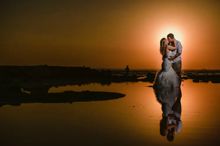 Bride and groom kissing at sunset, reflected in still water