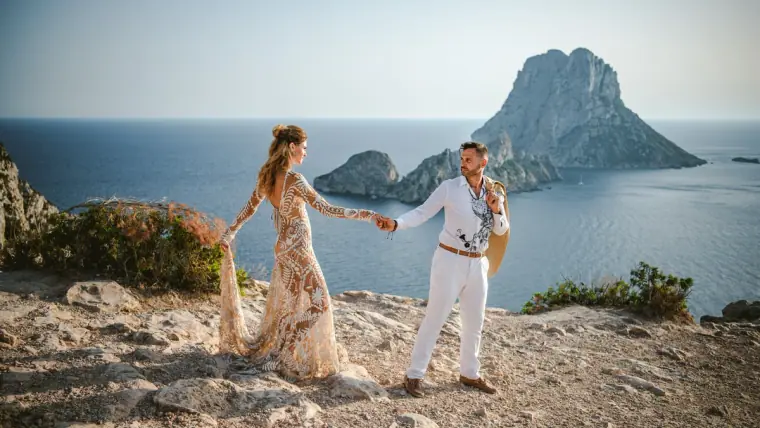 Bride and groom holding hands on a clifftop with a rocky island rising from the sea