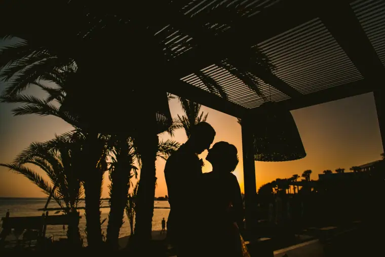 Silhouette of a couple kissing under palm-frond shade against an orange sunset
