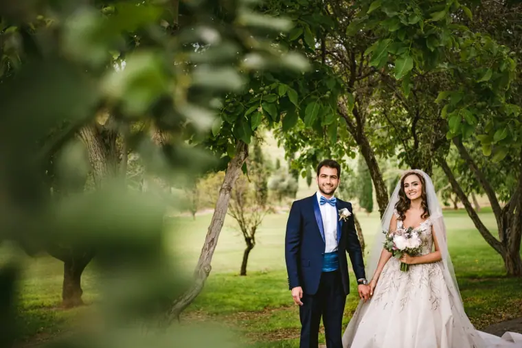 Groom in a navy suit and bride with a pink bouquet holding hands in a leafy orchard