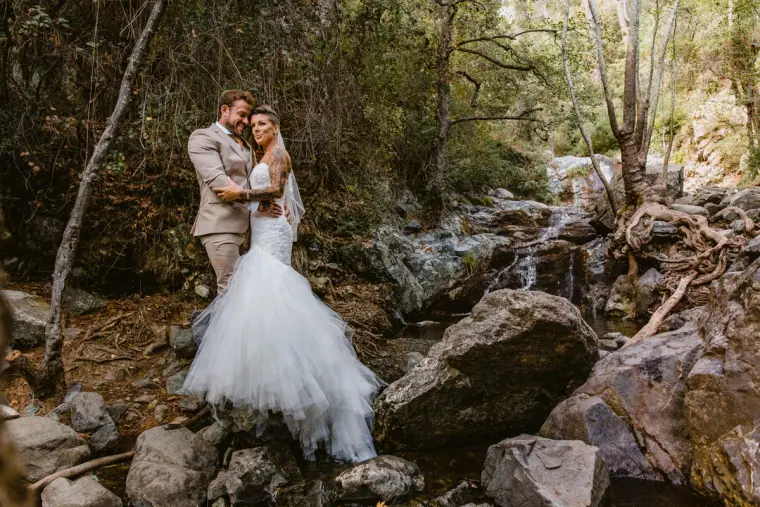 Bride in a mermaid gown and groom embracing among boulders by a woodland stream