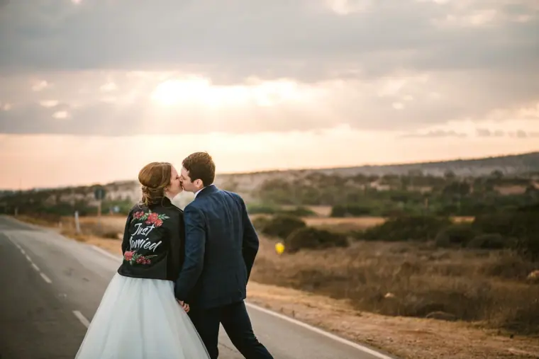 Bride in a custom painted jacket kissing the groom on a quiet country road at sunset